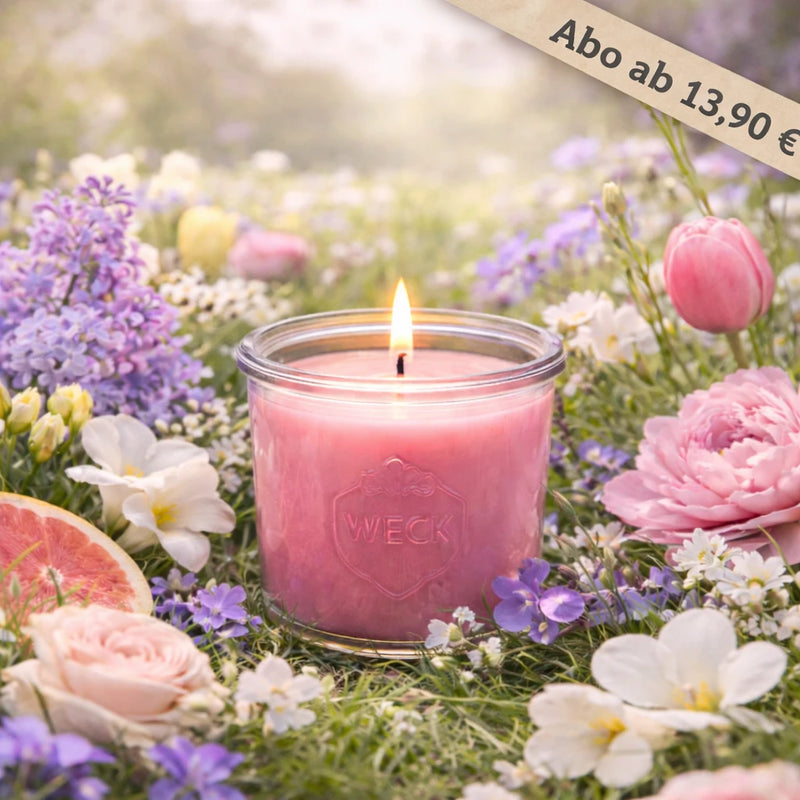 Brennende Duftkerze mit rosa Wachs im WECK-Glas, umgeben von einer bunten Blütenwiese mit Flieder, Tulpen, Veilchen, Pfingstrosen, Freesien, Rosen und einer Grapefruitscheibe – natürlicher Frühlingsmoment im Sonnenlicht.
