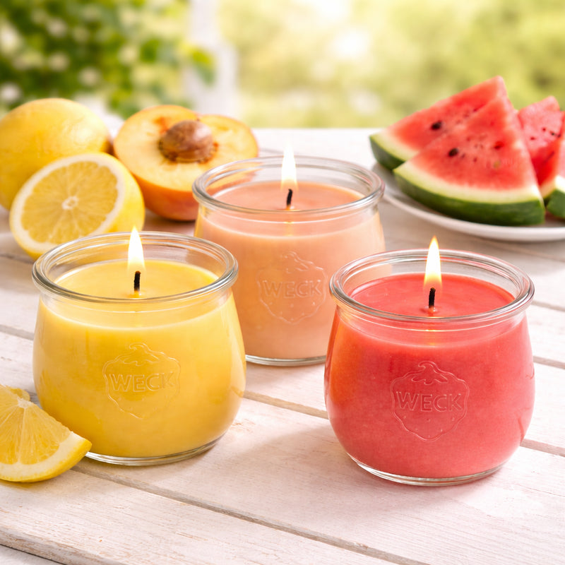 Drei Duftkerzen im WECK-Glas – in den Farben Zitronengelb, Pfirsich und Wassermelonenrot – brennen auf einem weißen Holztisch im Freien. Daneben liegen frische Zitronenscheiben, ein aufgeschnittener Pfirsich und ein Teller mit Wassermelonenscheiben. Sommerlich-leichte Stimmung mit natürlichen Farben.