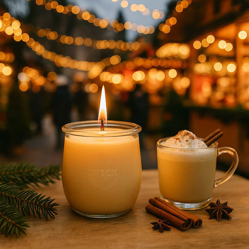 Brennende Duftkerze „Süßer Schwips“ mit cremefarbenem Wachs auf einem weihnachtlich dekorierten Tisch im Freien, umgeben von Lichtern und Tannenzweigen – stimmungsvolle Szene mit Eierpunsch-Flair auf dem Weihnachtsmarkt.