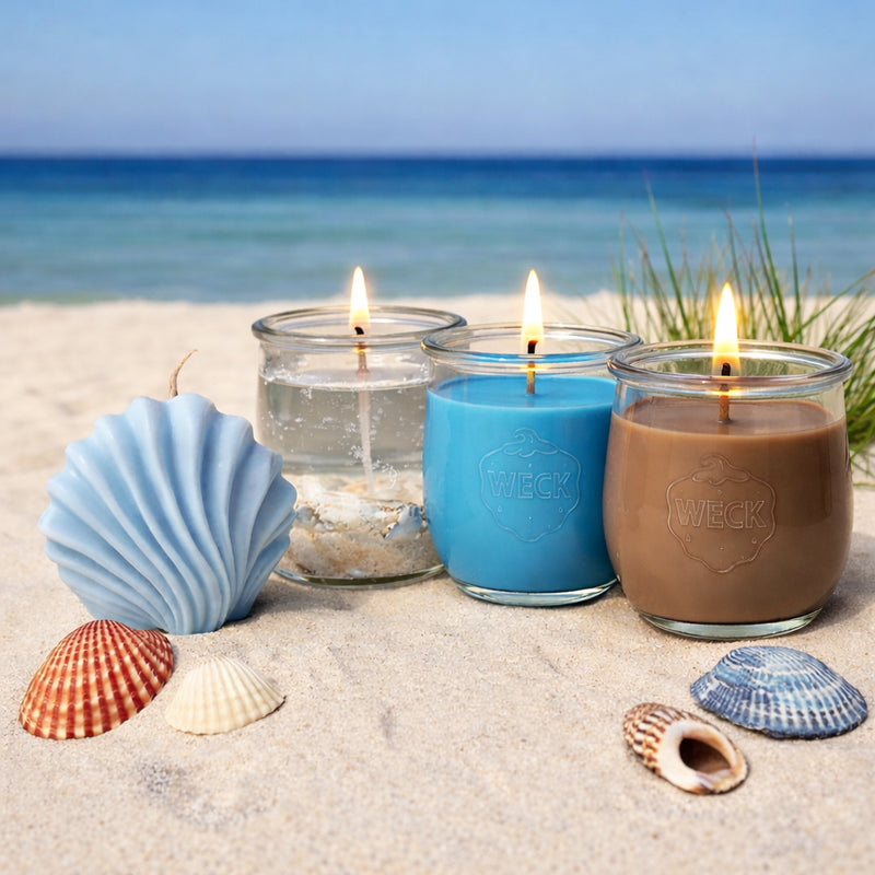 Vier verschiedene Duftkerzen auf Sand vor Meereskulisse: eine muschelförmige Deko-Kerze in Hellblau, eine transparente Gelkerze mit Muscheln, eine große blaue Duftkerze und eine große braune Duftkerze im WECK-Glas. Umgeben von echten Muscheln und Strandgras.