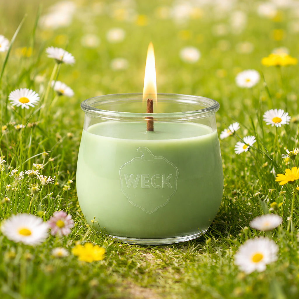 Pastellgrüne Duftkerze im WECK-Glas mit brennendem Docht auf einer blühenden grünen Frühlingswiese.