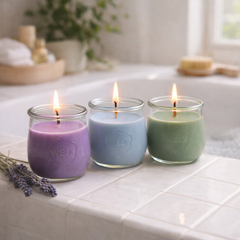 Drei brennende Duftkerzen im WECK-Glas stehen auf einer hellen Fliesenkante am Badewannenrand. Die Wachsfarben sind Lavendel, Hellblau und Salbeigrün. Im Hintergrund liegt ein Lavendelbund, weiches Tageslicht fällt durch das Fenster, Badutensilien sorgen für eine ruhige, entspannte Atmosphäre.