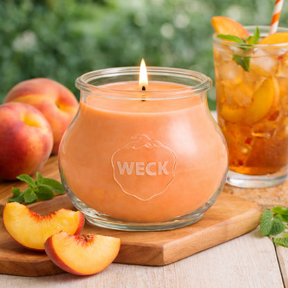 Pfirsichfarbene Duftkerze im runden Weckglas, brennend auf Holzplatte neben frischen Pfirsichen und einem Glas Eistee – stimmungsvolles Sommerbild im Freien