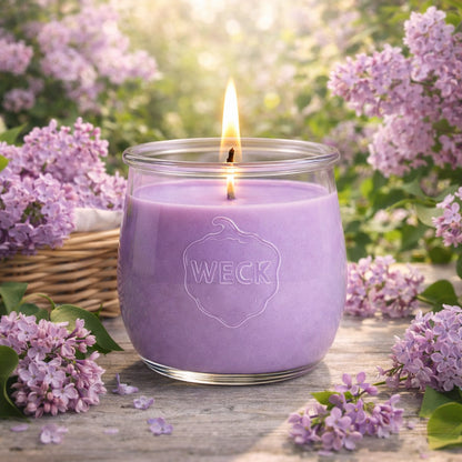 Fliederduftkerze im WECK-Glas, brennend auf Holztisch im Garten – umgeben von blühendem Flieder, Korbdeko und weichem Sonnenlicht – stimmungsvolle Frühlingsszene mit romantischem Flair.