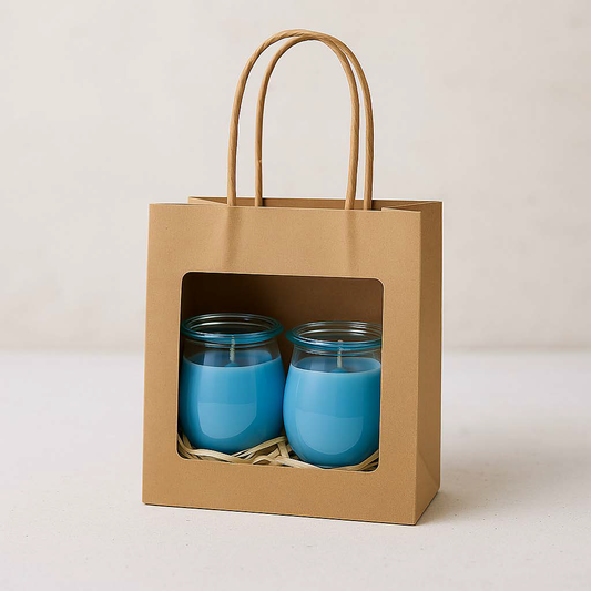 Zwei blaue Mini-Duftkerzen in einer Kraftpapiertüte mit Sichtfenster – Sorten Meeresbrise & Frische Wäsche, dekorativ verpackt mit Holzwolle.