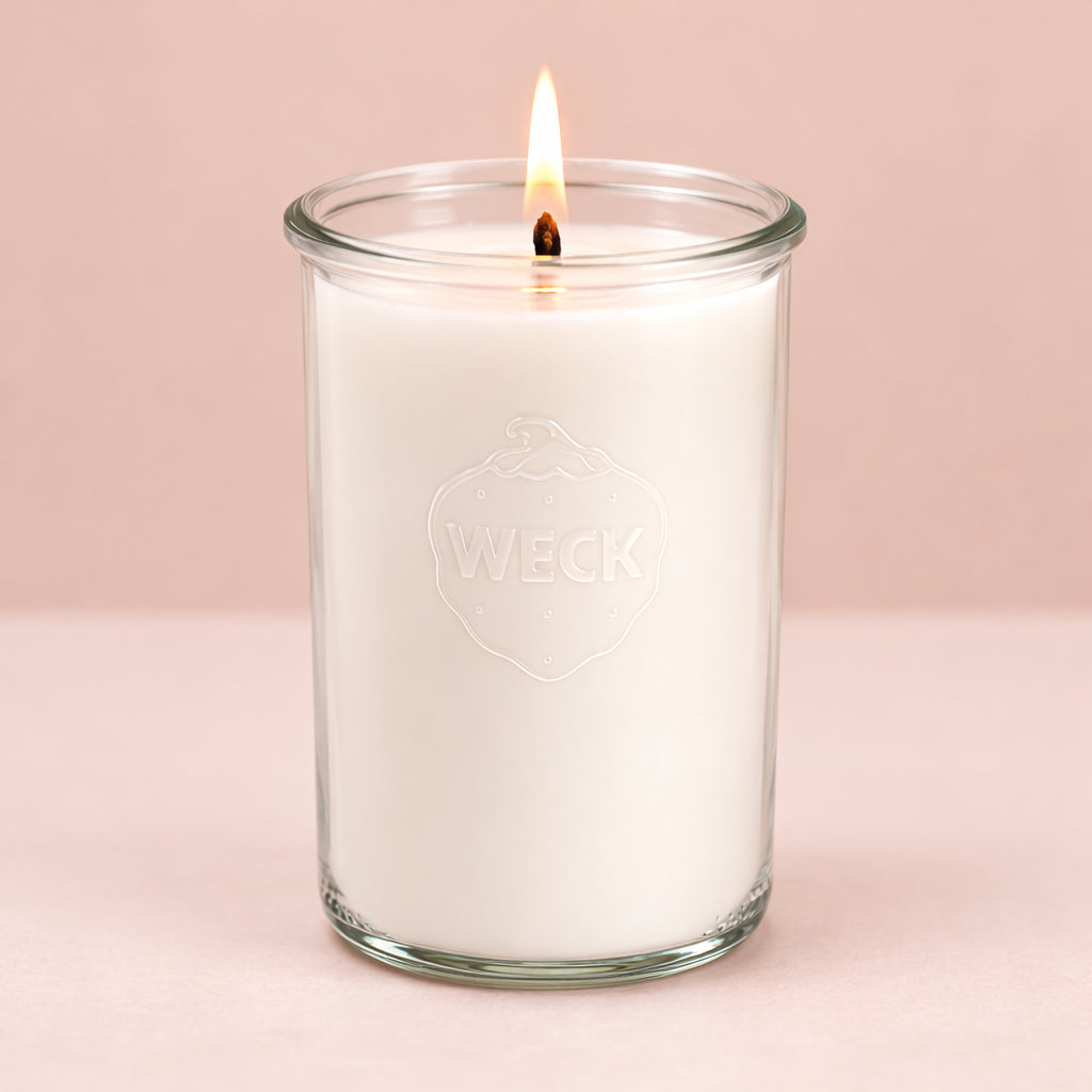 Hochformatiges Produktfoto einer weißen Duftkerze im hohen WECK-Glas, brennend, vor neutralem beigefarbenem Hintergrund. Die Kerze wirkt klar, schlicht und edel.