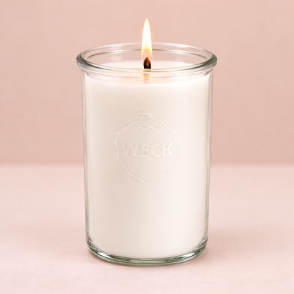 Hochformatiges Produktfoto einer weißen Duftkerze im hohen WECK-Glas, brennend, vor neutralem beigefarbenem Hintergrund. Die Kerze wirkt klar, schlicht und edel.