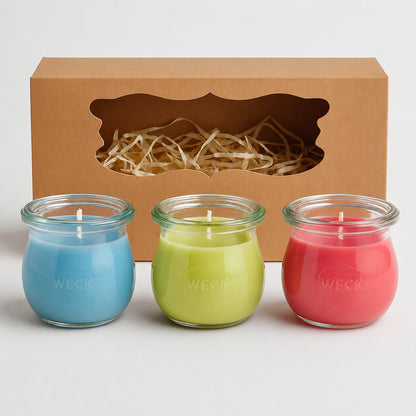 Drei Mini-Duftkerzen von CandleDeko in Blau, Grün und Pink vor Kraftkarton-Geschenkbox mit Sichtfenster