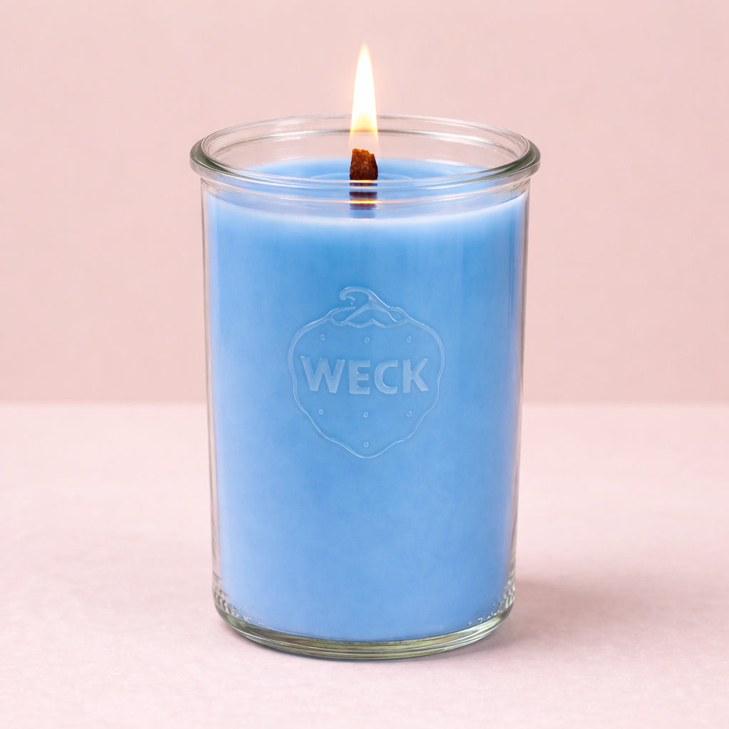 Brennende Duftkerze Meeresbrise im WECK-Glas auf Holzbrett mit Muscheln und Treibholz vor Dünen und Meer im Hintergrund.