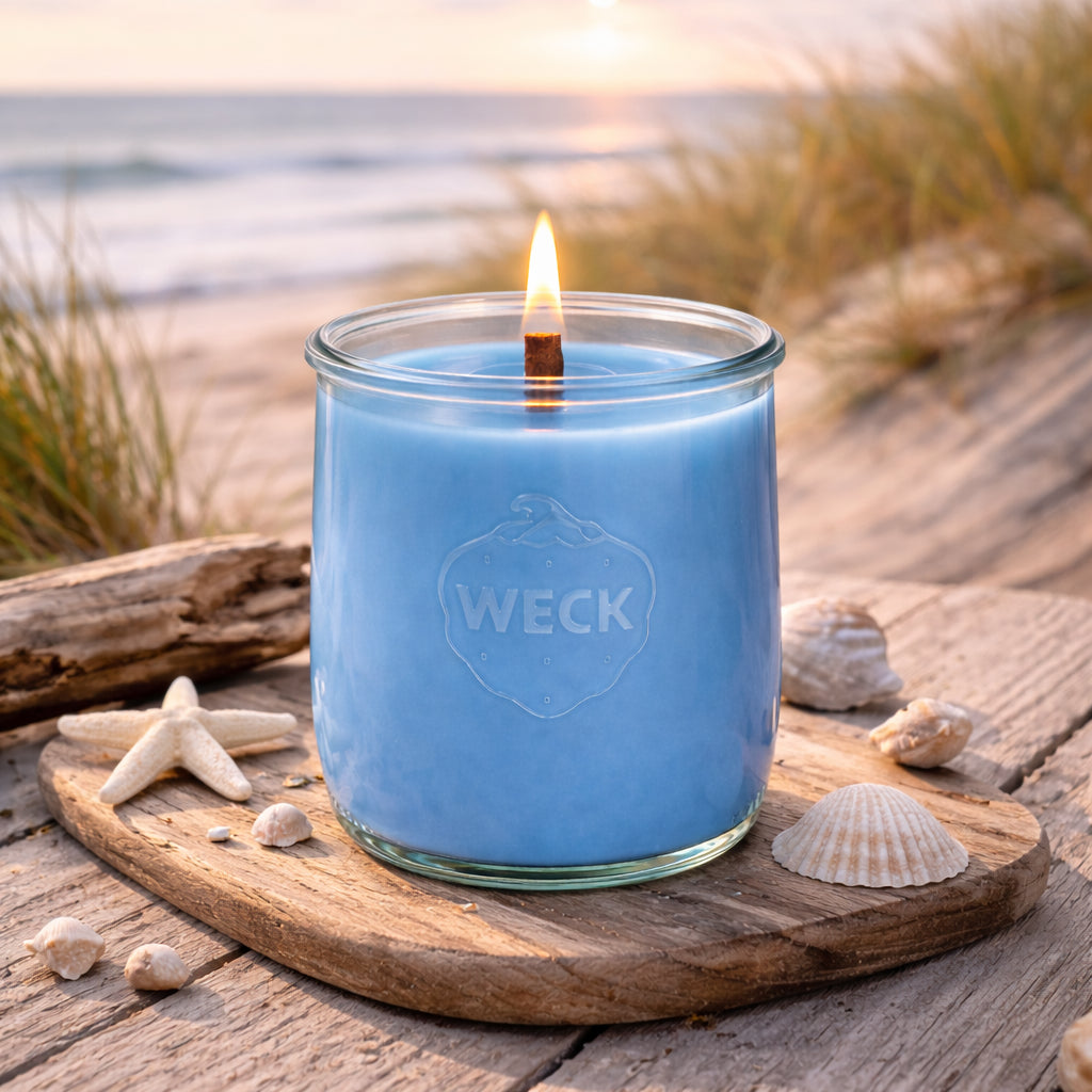 Duftkerze Meeresbrise im großen WECK-Glas mit blauem Wachs auf Holzbrett am Strand, Muscheln und Treibholz im warmen Abendlicht.