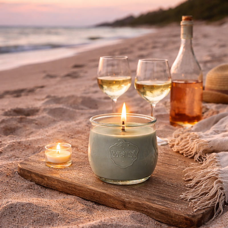 Romantische Szene am Strand mit brennender eukalyptusgrauer Duftkerze im WECK-Glas, zwei Gläsern Wein, Roséflasche und Stranddecke bei Sonnenuntergang