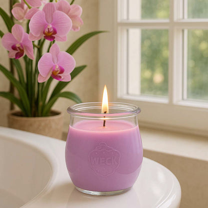 Duftkerze „Orchidee“ brennt am Rand einer weißen Badewanne, mit rosa Orchideen und Fenster im Hintergrund – helles, entspannendes Frühlingsambiente.