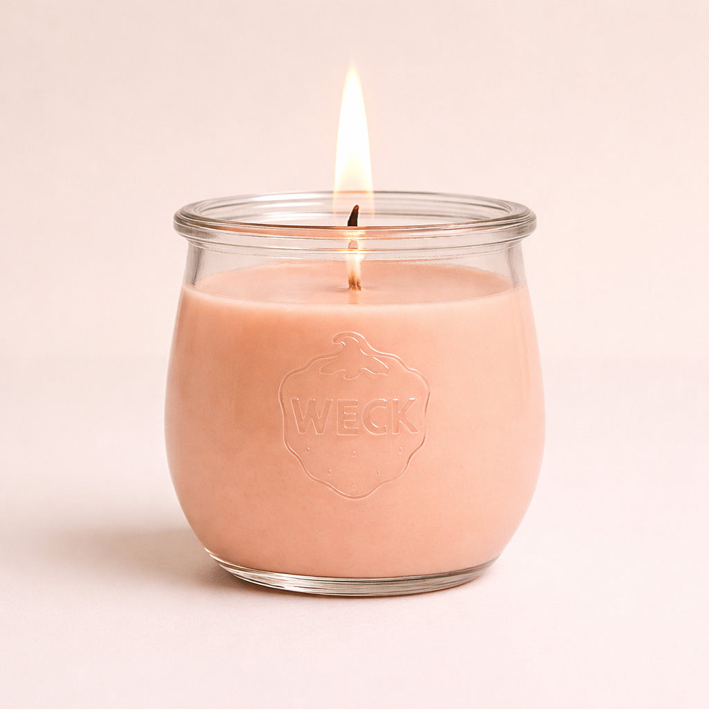 Brennende Duftkerze in sanftem Pfirsichton im WECK-Glas mit geprägtem Logo, vor warmem Hintergrund mit weichem Schatten – klare Studioaufnahme.