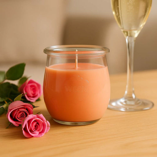 Große pfirsichfarbene Duftkerze im Glas, brennend in einem eleganten Ambiente mit Rosen und Schaumwein – vermittelt sommerlich-festliche Stimmung