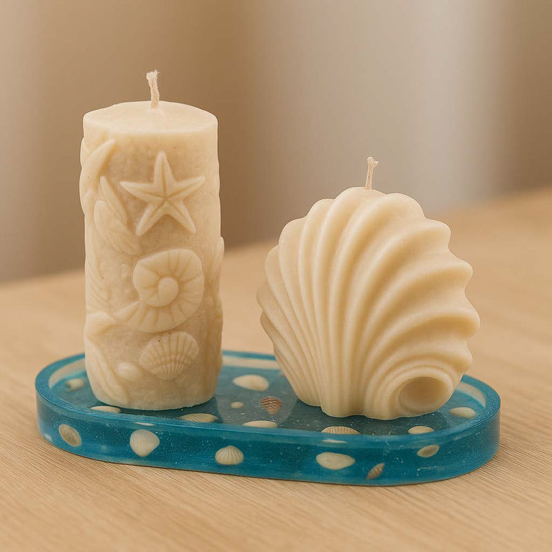 Zwei beige Kerzen in Meeresform – eine Muschelkerze und eine Stumpenkerze mit Seestern- und Muscheldetails – stehen auf einem sechseckigen, kleegrünen Epoxidharz-Untersetzer. Die Komposition vermittelt maritimes Flair und stilvolle Natürlichkeit.