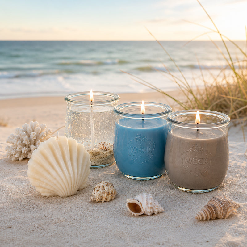 Vier verschiedene Kerzen auf Sand vor Meereskulisse: eine muschelförmige Deko-Kerze in Hellblau, eine transparente Gelkerze mit Muscheln, eine große blaue Duftkerze und eine große braune Duftkerze im WECK-Glas. Umgeben von echten Muscheln und Strandgras.