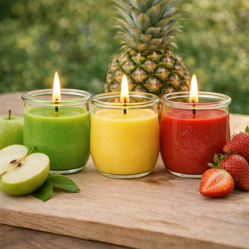 Drei brennende Duftkerzen im WECK-Glas in den Farben Grün, Gelb und Rot stehen nebeneinander auf einem Holztisch. Neben den Kerzen liegen frische Früchte: Apfel, Ananas und Erdbeeren. Im Hintergrund unscharfer, grüner Naturhintergrund.