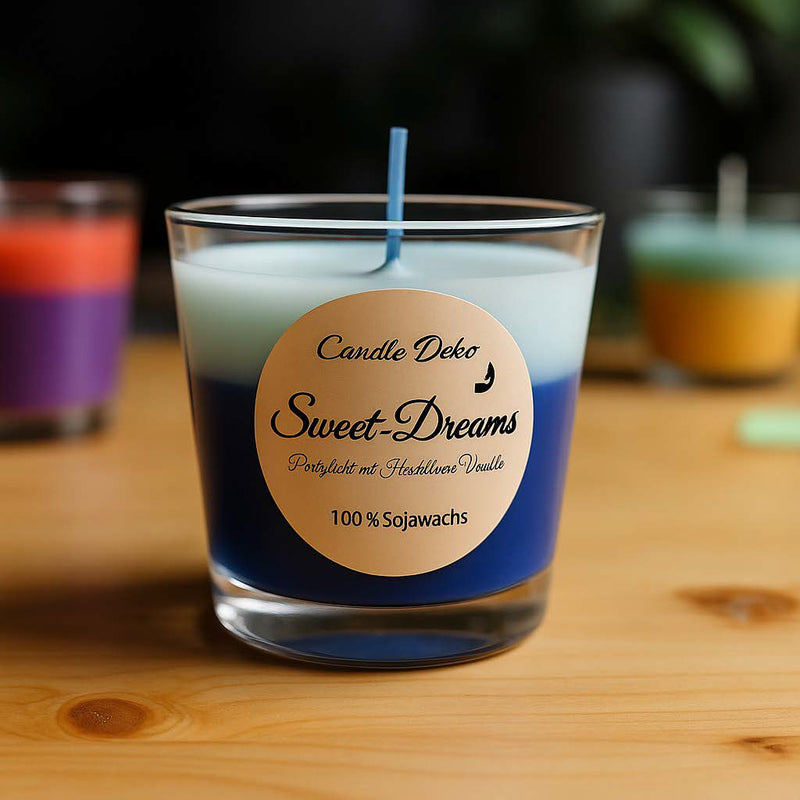 Zweifarbige Duftkerze „Sweet-Dreams“ in einem klaren Glas mit Etikett, bestehend aus dunkelblauer und pastellblauer Wachsschicht, auf einem Holztisch. Im Hintergrund weitere farbige Kerzen und unscharfe Pflanzen – warmes, natürliches Licht erzeugt eine gemütliche Stimmung.