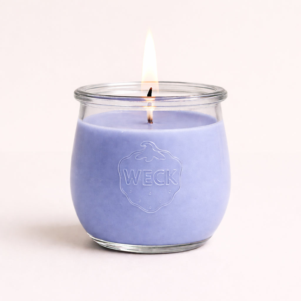 Brennende Duftkerze in lila-blauem Wachs im WECK-Glas mit geprägtem Logo, vor neutralem Hintergrund mit weichem Schatten – ruhige, hochwertige Studioaufnahm