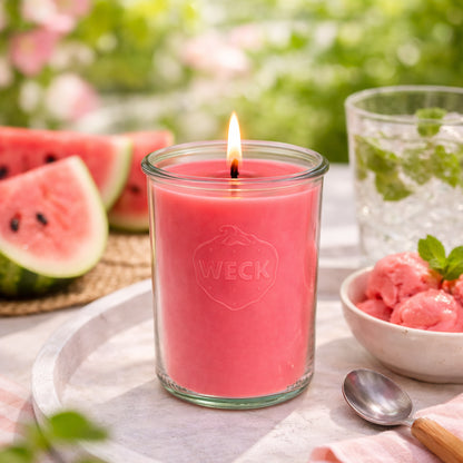 Brennende rote Duftkerze im WECK-Glas auf Naturmatte, umgeben von frischen Wassermelonenstücken, Minzblättern und einem Glas Wassermelonendrink – sommerliche Szene für die Duftkerze Wassermelonensorbet.