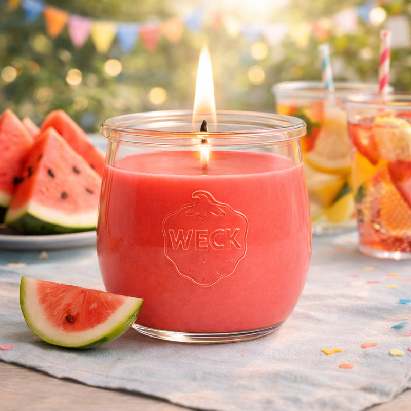 Wassermelonensorbet-Duftkerze im WECK-Glas, brennend auf sommerlich gedecktem Tisch mit Melonenstücken, bunten Getränken und Partygirlanden – fröhliche Outdoor-Stimmung im Abendlicht.