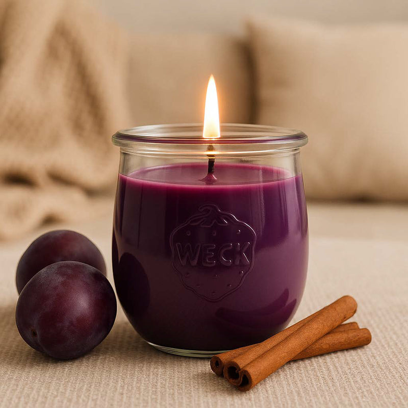 Duftkerze Winterpflaume im WECK-Glas, angezündet, umgeben von frischen Pflaumen und Zimtstangen auf hellem Stoff – stimmungsvolle Winterdeko in warmen Naturtönen.