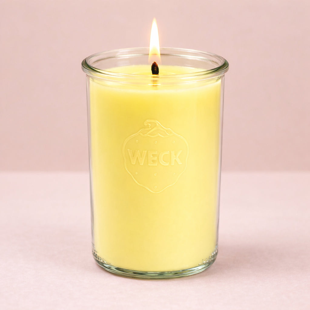 Eine hohe Duftkerze im WECK-Glas mit zitronengelbem Wachs brennt ruhig vor einem beigefarbenen, weichen Hintergrund. Die Flamme steht ruhig, das Design ist schlicht und klar