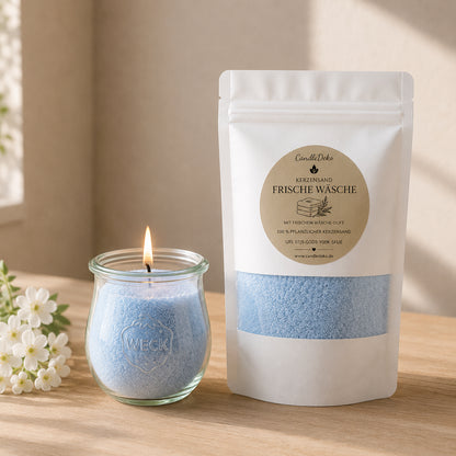 Ein weißer CandleDeko Nachfüllbeutel mit hellblauem Duft-Kerzensand Frische Wäsche neben einer brennenden Kerze im bauchigen WECK-Glas auf einem Holztisch mit weißen Blüten.