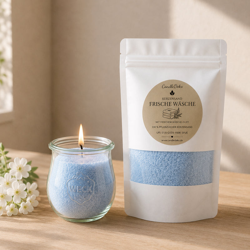 Ein weißer CandleDeko Nachfüllbeutel mit hellblauem Duft-Kerzensand Frische Wäsche neben einer brennenden Kerze im bauchigen WECK-Glas auf einem Holztisch mit weißen Blüten.