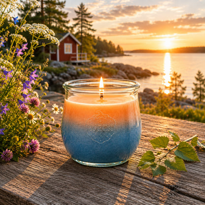 Große Midsommer Duftkerze aus Kokoswachs im WECK-Tulpenglas neben auf einem Holztisch am skandinavischen See. Die warmen Farben der Mitternachtssonne spiegeln sich im ruhigen Wasser und schaffen die besondere Stimmung eines nordischen Sommerabends.