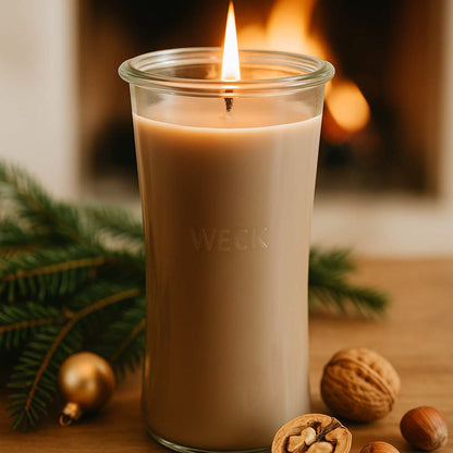Nussknacker XXL Duftkerze aus Kokoswachs im großen WECK-Glas von CandleDeko, braun und brennend auf Holztisch neben Nüssen vor Kaminfeuer.